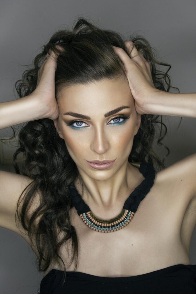 Close-up portrait of a brunette woman with striking makeup and necklace, hands on head.